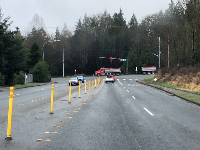A three-way intersection of four lane roads coming to a traffic light surrounded by trees. There is too much road capacity to keep those trees.