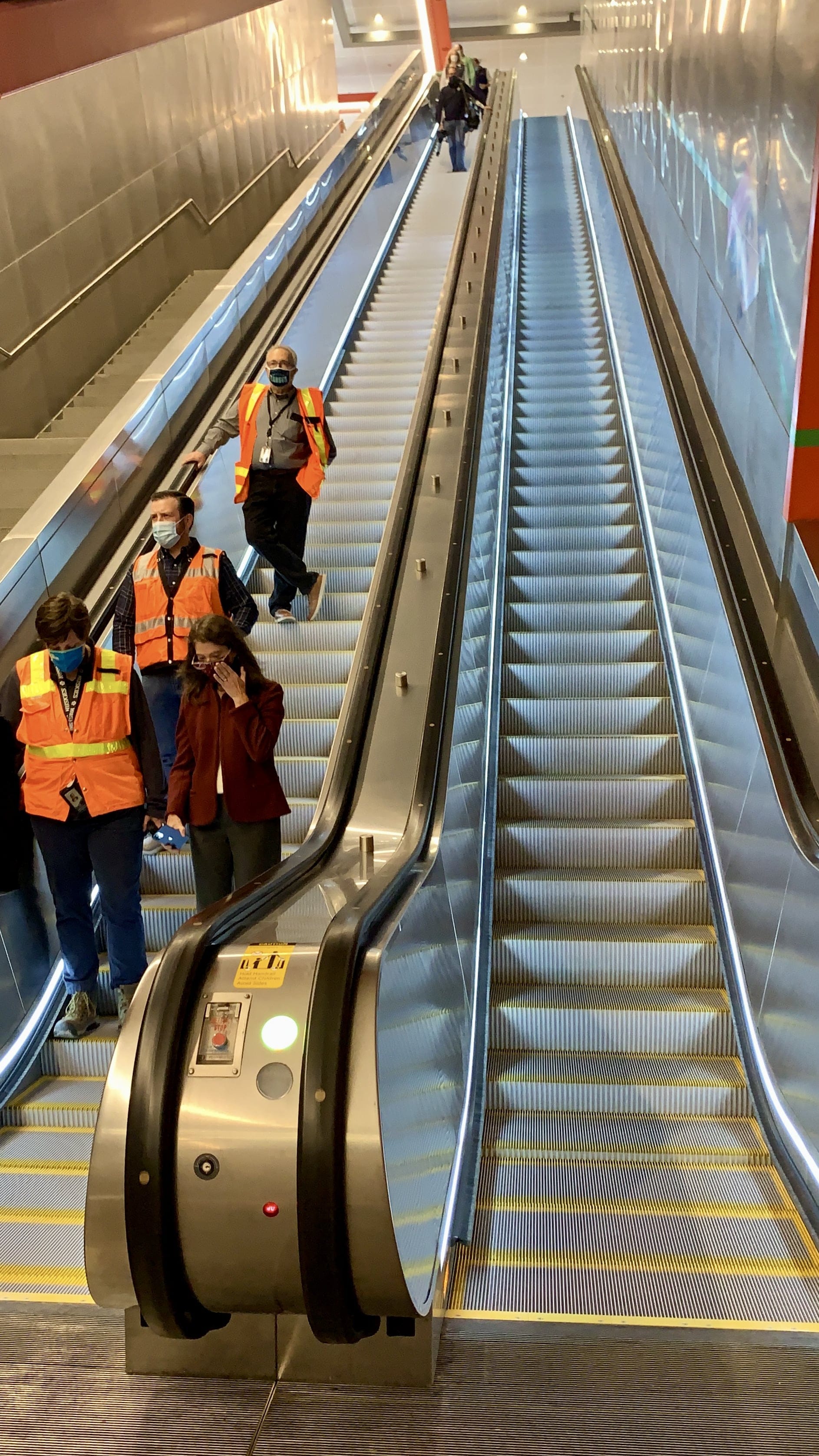 A photo looks up at a long, steep set of escalators that lead into the station. People wearing orange safety vests and a woman in a red coat ride the escalator. 