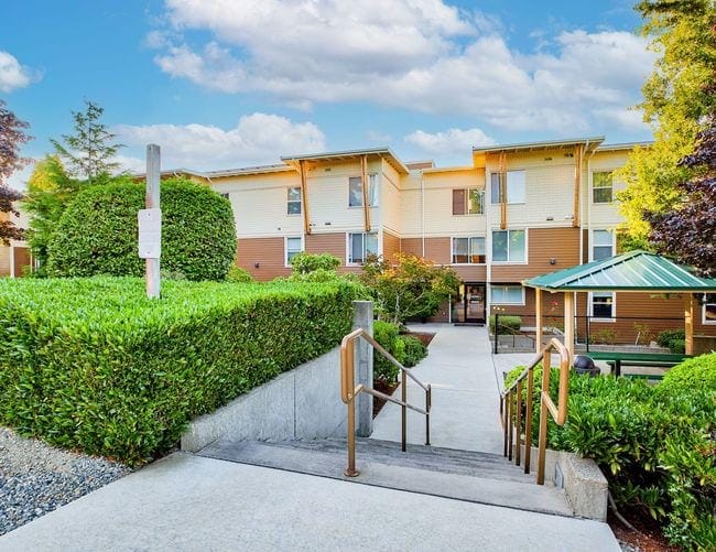 A photo of Burien Haus apartments, a tan and cream colored three story apartment building surrounded by green bushes and trees.