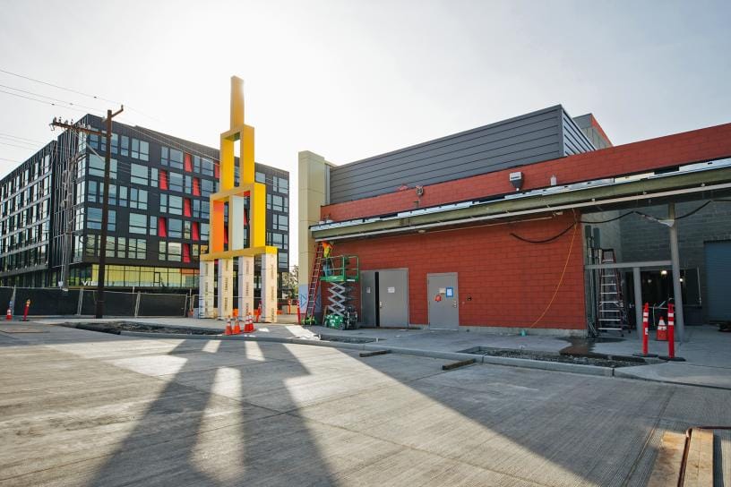 A photo showing a large plaza with a statue of interlocking yellow squares, and light rail station under construction. 