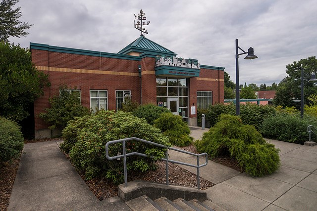 A photo of a red brick building with a sidewalk entrance and shrubs. 