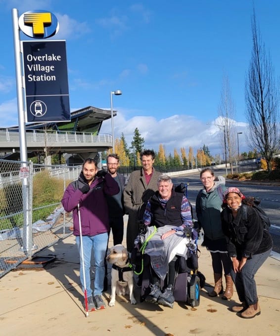A photo shows a man standing in a purple coat with a white cane, near to two men standing in grey coats behind a man seated in a wheelchair with a seeing eye dog. Two women also in coats are crouching next to him. 