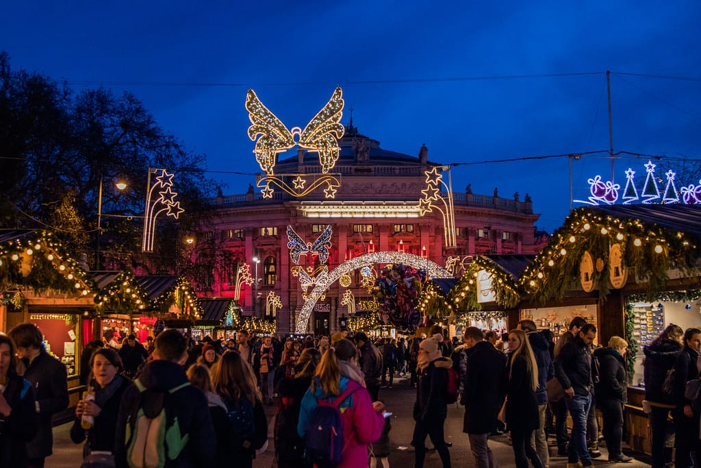 A crowd of people before a historic building illuminated red, wooden kiosks peeling items, and holiday lights.