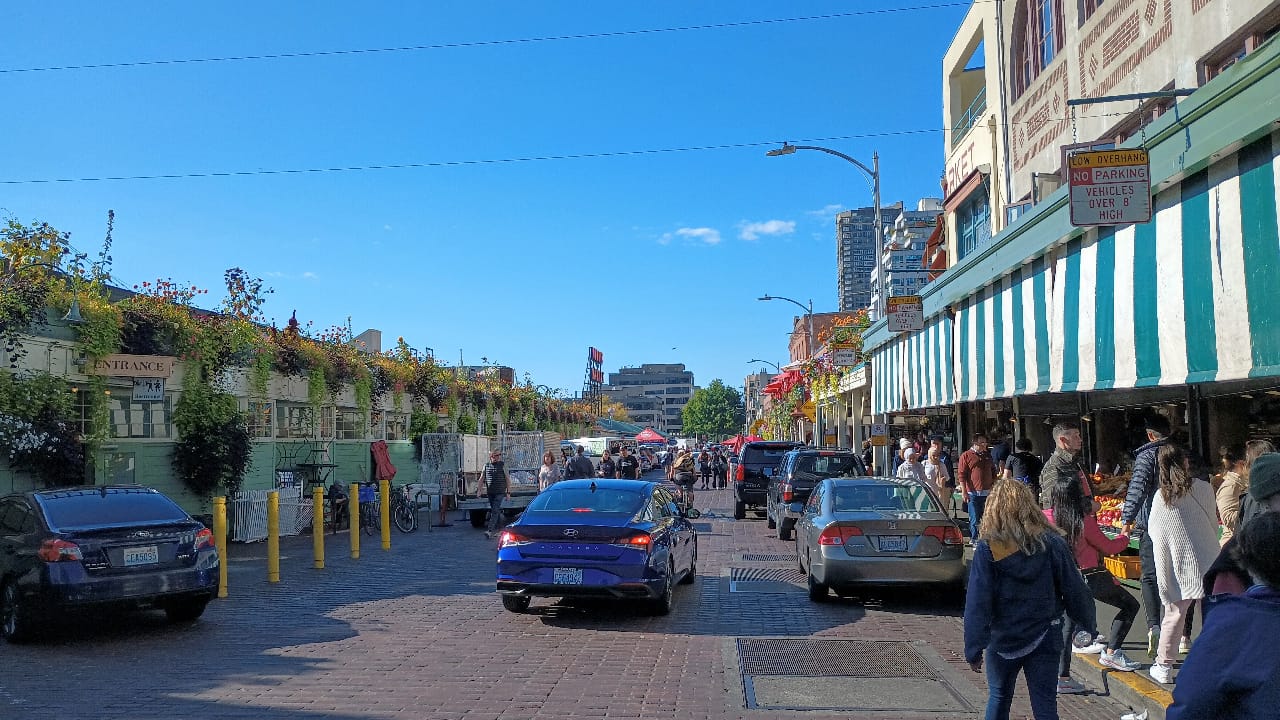 
                     Cars parked and motoring past shops at Pike Place Market.
                     
