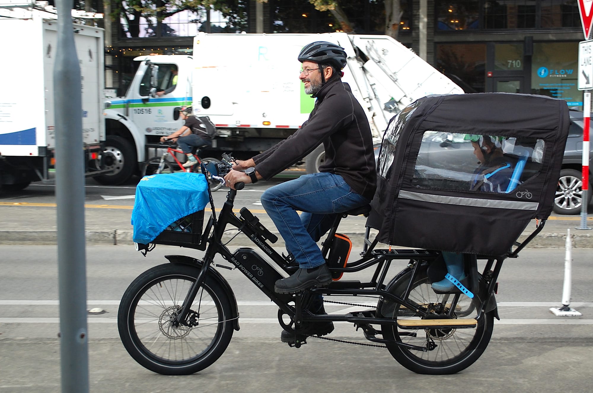 An e-biker with cargo above the front wheel and a child carrier with weather protection over the back wheel.