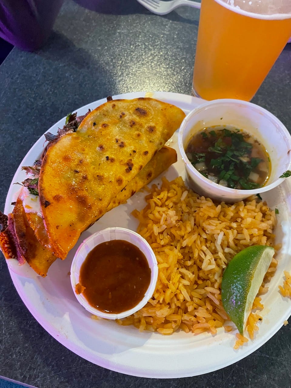 Two tacos with rice and dipping sauce with a remarkably pale beer next to them.