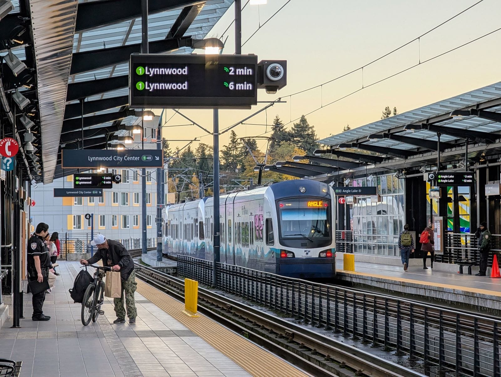 
                     A southbound train departs with an apartment building in the background. A man rolls a bike on the platform and security guar
                     