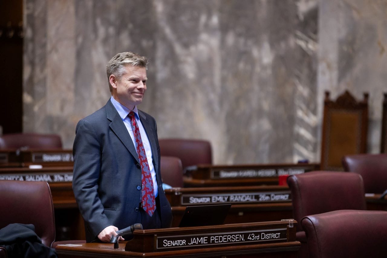 Pedersen stands in the senator chambers in a suit with a marble wall in the background.