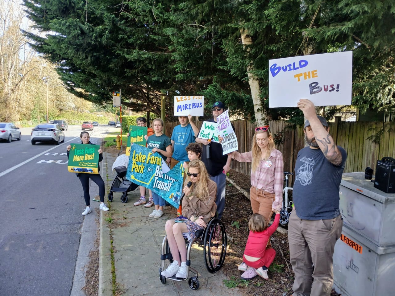 Cecilia hold a microphone and sits in a power wheelchair. Transit advocates stand behind her on the sidewalk holding signs, including seven adults, two kids, and a baby.