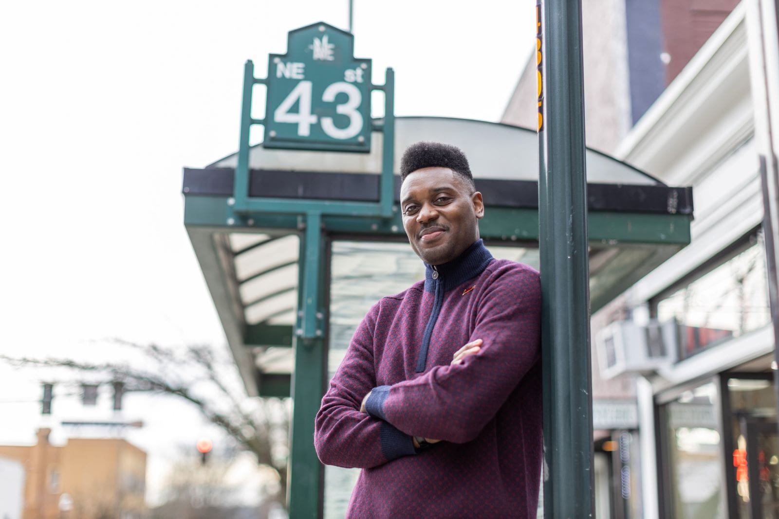 Shaun stands in front of a bus stop and leans on a street pole with a NE 43rd St sign in the background. Shaun is a  millennial and Black man with his arms crossed.