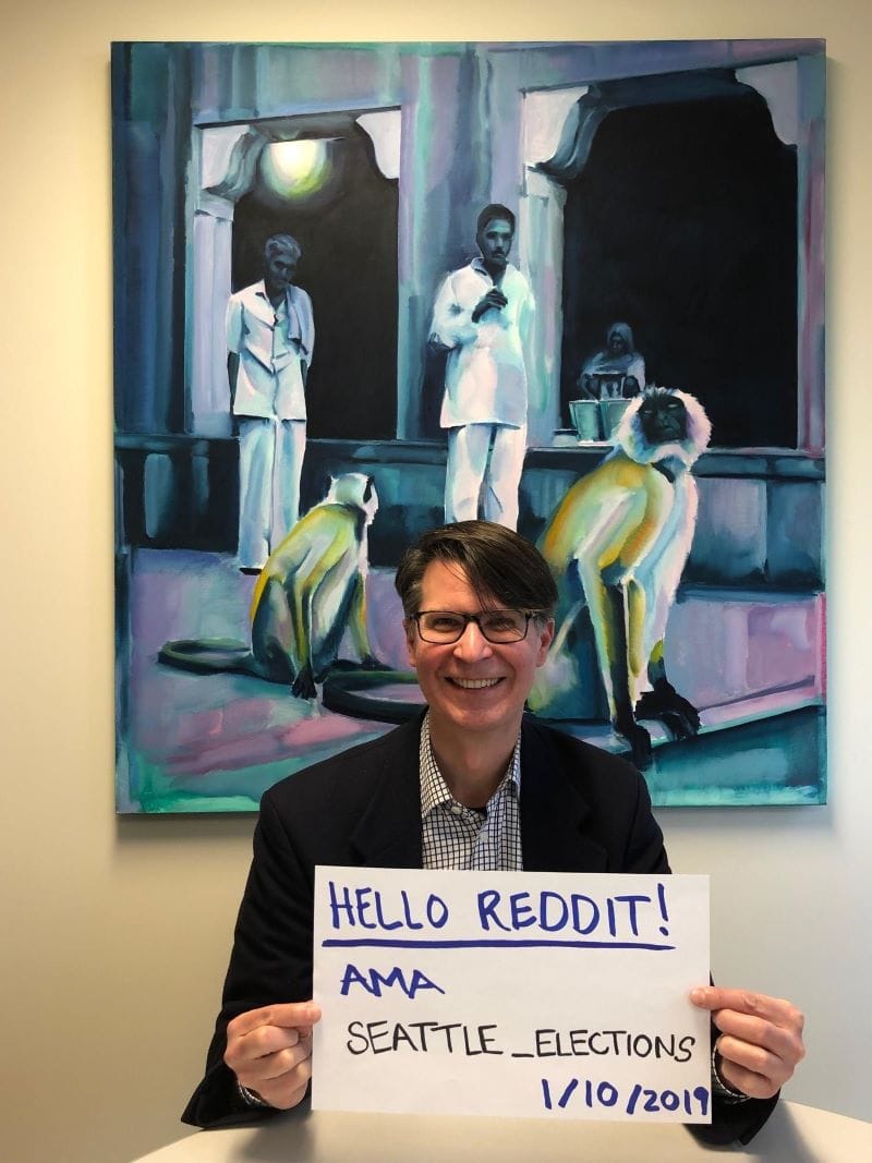Wayne Barnett smiles and hold a sign that says "Hello Reddit! AMA Seattle Elections 1/19/2019." A paining with two lower primates and two humans is the background.