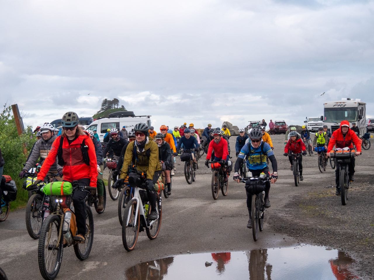 A big crowd of dozens of bike riders with an RV in the background an overcast sky.