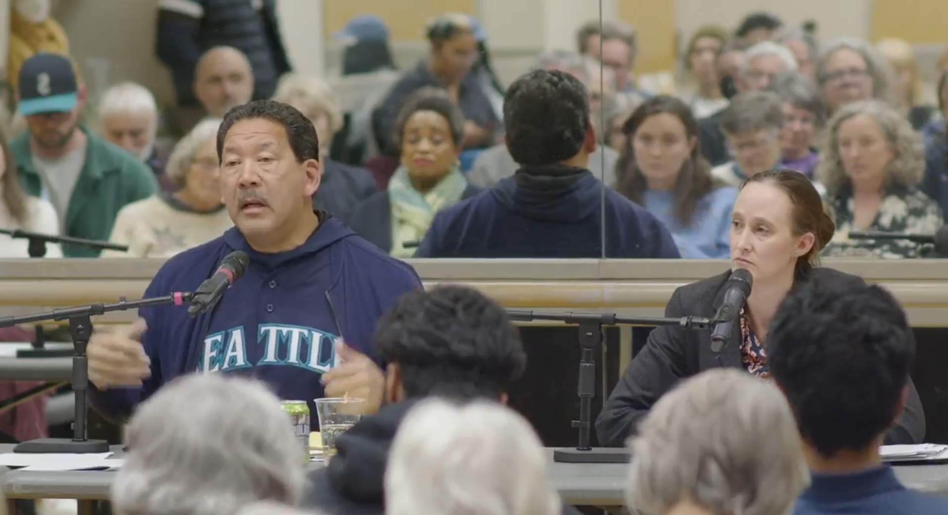 Mayor Harrell gestures with his hand as he speaks and wears a Mariners jersey. Wilson is seated just to the right. A seated crowd listens in the background.