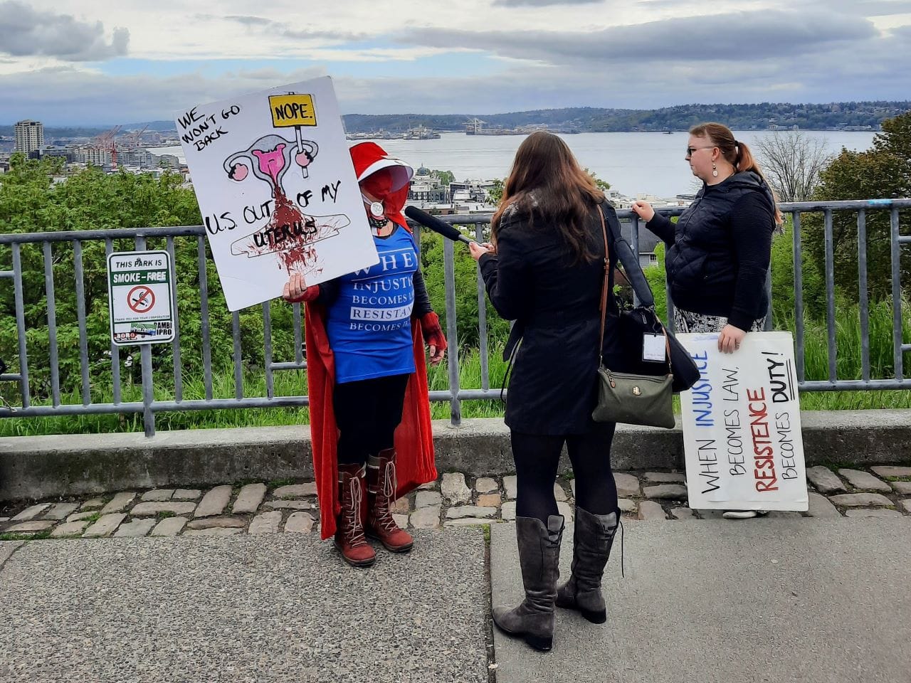 A woman wearing a bright red cape in the style of Handmaids Tale holds a sign showing a cartoon uterus and saying "We won't go back. Nope. U.S. out my uterus." She is being interviewed by a reporter at Kerry Park.