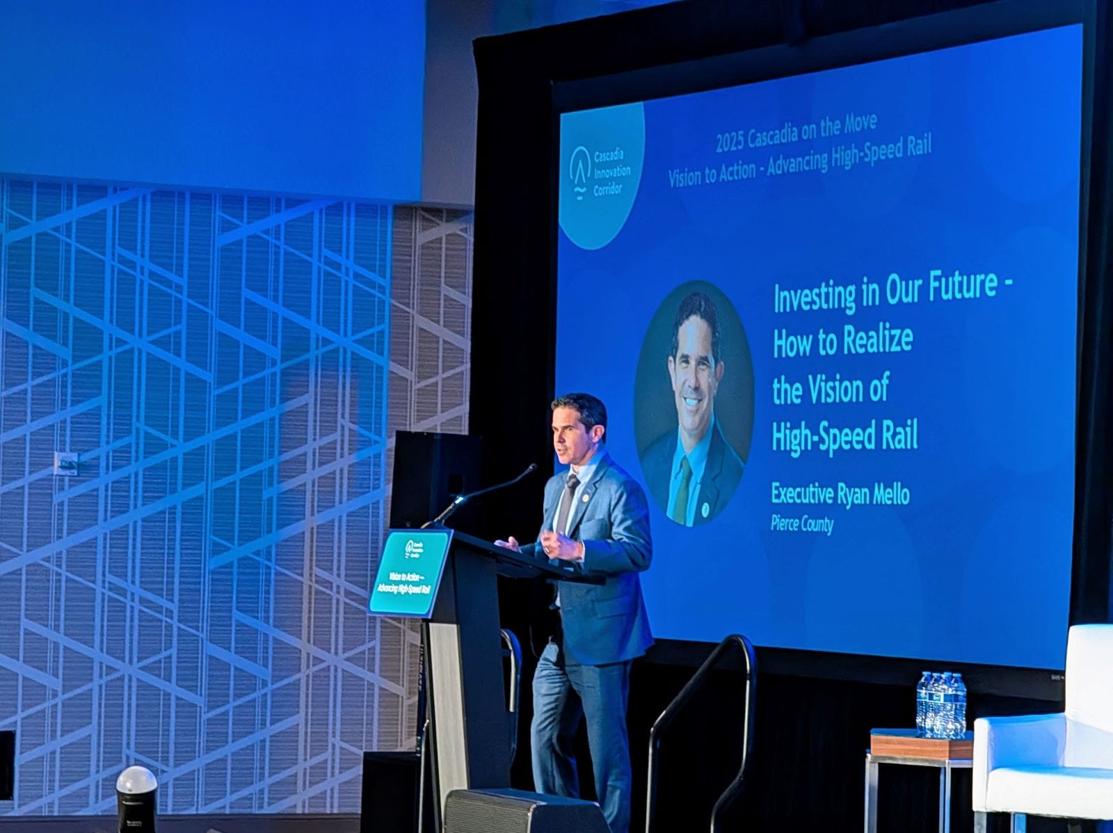 Mello wears a gray suit and stands at a lectern on stage. The slide on the projector behind him says "Investing in our future - how to realize the vision of high speed rail"