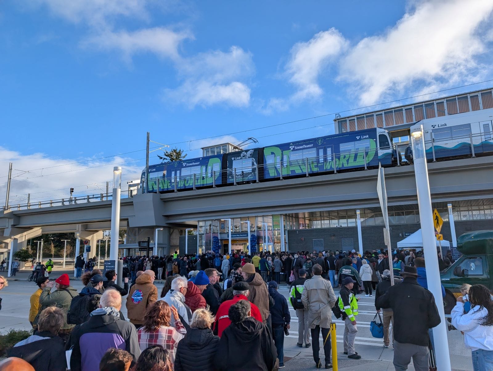 A line of people approach Federal Way Downtown Station, with a train waiting at the elevated platform.