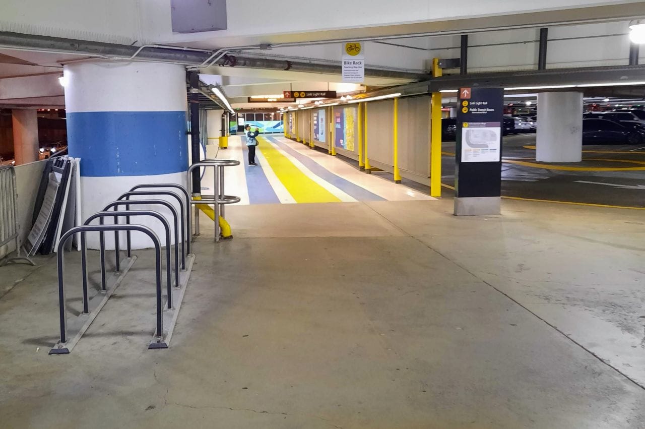 A line of staple bike racks, an elevator bank, and yellow and blue stripes guiding the path to light rail greet passengers as they enter the parking garage.